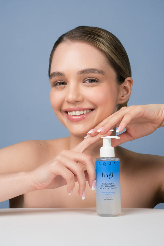 uśmiechnięta kobieta, przed którą stoi Hagi Aqua Zone Łagodny Żel-Galaretka do Mycia Twarzy 150ml, kobieta obiema dłońmi dotyka produktu