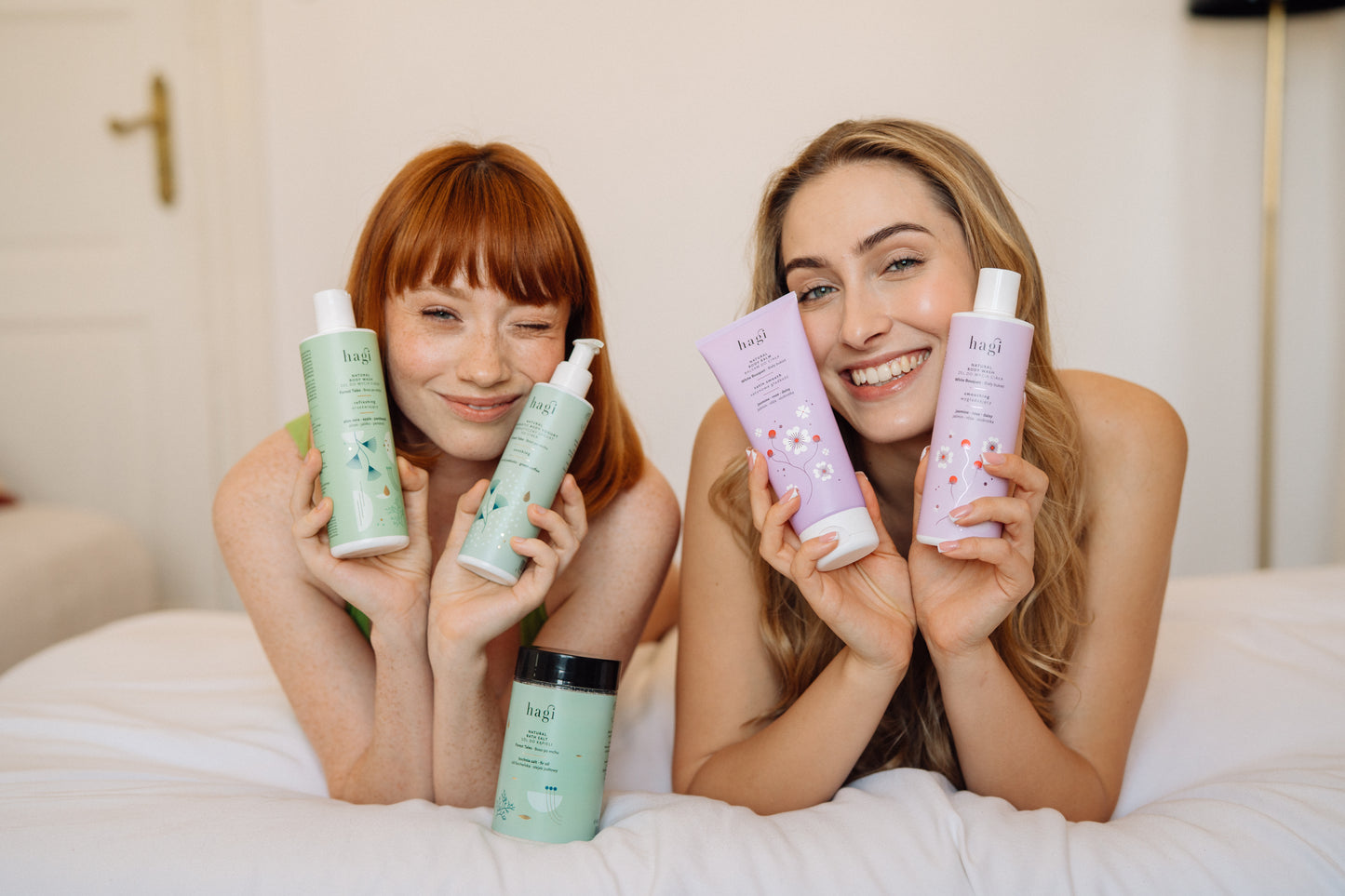 dwie uśmiechnięte kobiety leżąc na łóżku przodem w kierunku aparatu trzymają Hagi Naturalny Żel do Mycia Ciała Boso po Mchu 300 ml, Hagi Naturalny Żel do Mycia Ciała Biały oraz Hagi Naturalny Balsam do Ciała Biały Bukiet 200 ml  