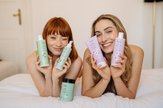 dwie uśmiechnięte kobiety leżąc na łóżku przodem w kierunku aparatu trzymają Hagi Naturalny Żel do Mycia Ciała Boso po Mchu 300 ml, Hagi Naturalny Żel do Mycia Ciała Biały oraz Hagi Naturalny Balsam do Ciała Biały Bukiet 200 ml  